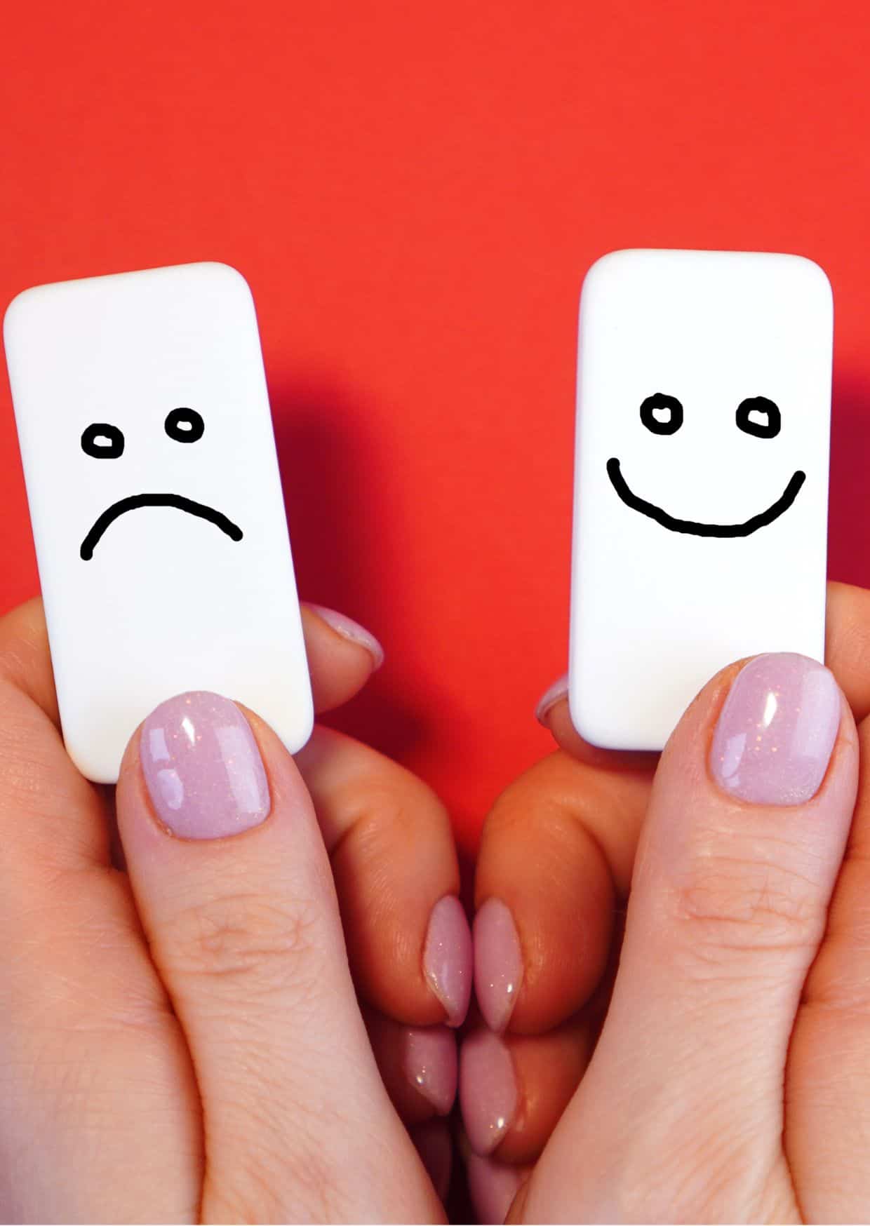 Zwei Hände mit pinkem Nagellack halten zwei weiße Kacheln nebeneinander. Auf einer Kachel ist ein handgezeichnetes trauriges Gesicht, auf der anderen ein fröhliches Gesicht.