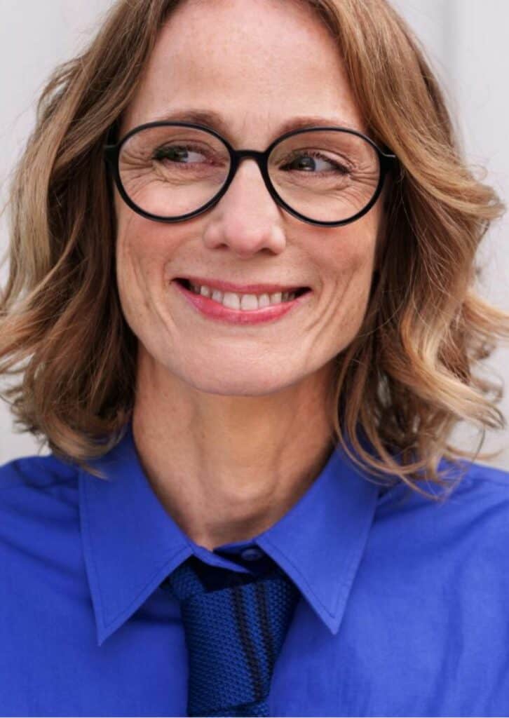 Porträt einer Frau mit blonden Haaren, großer Brille und blauem Hemd
