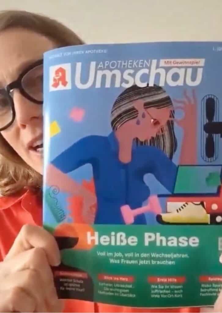 Frau mit großer Brille hält Zeitschrift in die Kamera.