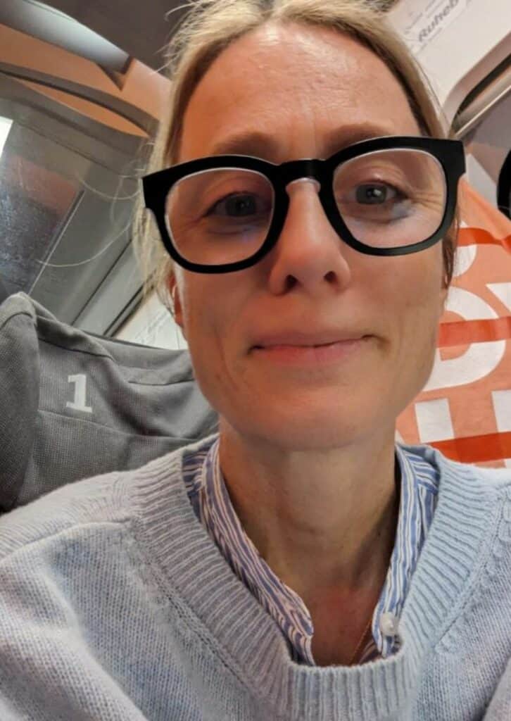 Ein Selfie einer Frau mit großen schwarzen Brillen und blonden Haaren, die einen hellblauen Pullover und eine gestreifte Bluse trägt. Sie sitzt in einem modernen Verkehrsmittel, vermutlich einem Zug, auf einem Sitzplatz mit der Nummer "1".