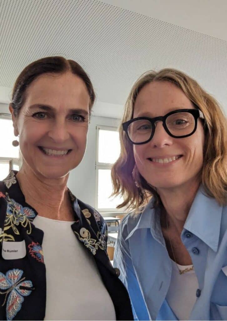 Ein Selfie von zwei Frauen, die lächelnd in die Kamera schauen. Die Frau links trägt eine Jacke mit buntem Blumenmuster und ein Namensschild mit der Aufschrift "Andrea Rumler". Die Frau rechts trägt eine Brille und ein hellblaues Hemd. Im Hintergrund befindet sich ein heller Raum mit Fenstern und Tischen.