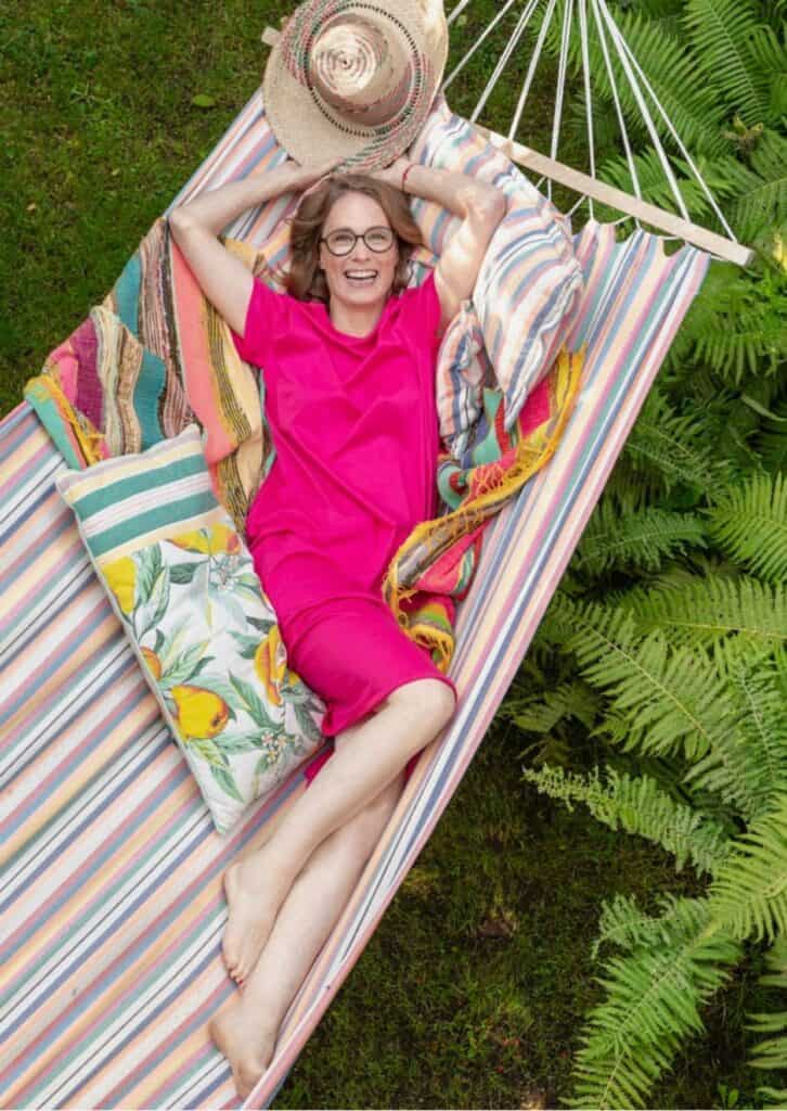Eine Frau liegt entspannt in einer bunt gestreiften Hängematte, umgeben von grünen Farnen. Sie trägt ein pinkfarbenes Kleid und eine Brille, und sie lächelt glücklich in die Kamera. Neben ihr liegen bunte Kissen und eine gestreifte Decke. Ein Strohhut liegt über ihrem Kopf.