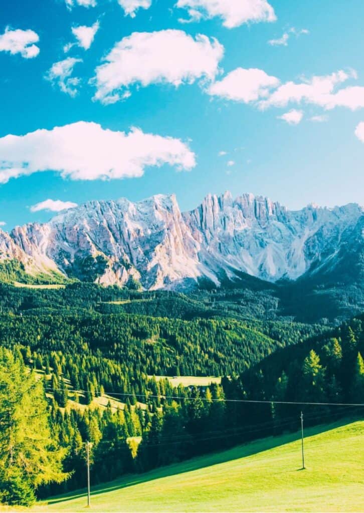 Eine malerische Landschaft mit hohen Bergen im Hintergrund, die von einem klaren blauen Himmel mit wenigen Wolken überragt werden. Die Berge haben schroffe, schneebedeckte Gipfel, während der Vordergrund von dichten Wäldern und grünen Wiesen geprägt ist. Das Sonnenlicht fällt hell auf die Szene und hebt die lebhaften Farben der Natur hervor.