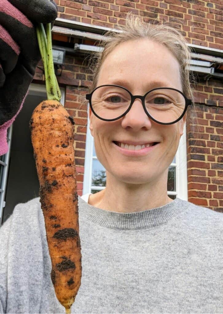 Eine Frau mit Brille und einem grauen Pullover hält lächelnd eine frisch geerntete, noch erdige Karotte in die Kamera. Im Hintergrund ist eine Backsteinwand zu sehen, die zu einem Haus gehört.