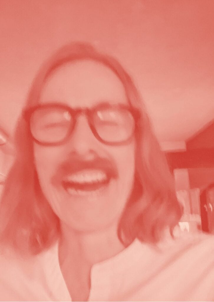 Ein verschwommenes Bild einer lachenden Person mit Brille und einem Schnurrbart. Das Bild ist in einem rötlichen Farbton gehalten, wodurch es eine künstlerische, abstrakte Wirkung hat.