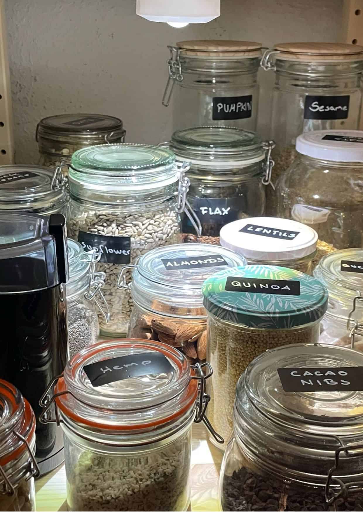 Glasgefäße in einem Vorratsschrank, ordentlich beschriftet mit verschiedenen Samen, Nüssen und Hülsenfrüchten. Zu sehen sind unter anderem Gläser mit Sonnenblumenkernen, Mandeln, Quinoa, Linsen, Kakaonibs, Hanfsamen, Leinsamen, Sesam und Kürbiskernen, beleuchtet von einer kleinen Lampe.