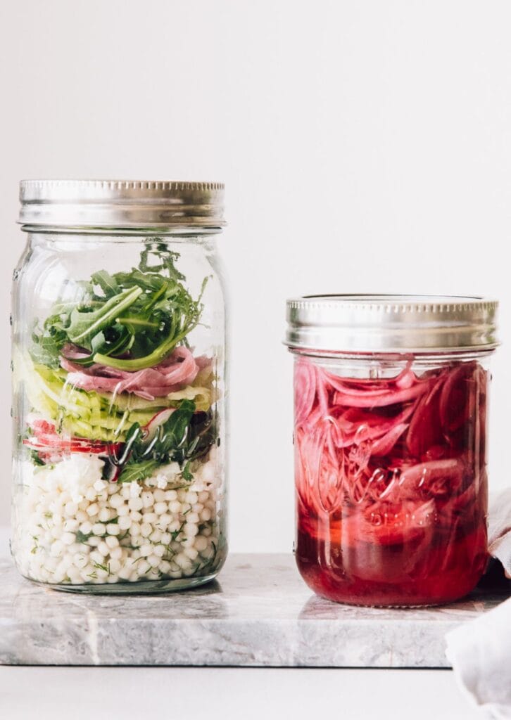 Zwei Einmachgläser mit fermentiertem Gemüse und frischen Zutaten auf Marmorplatte – gesunde, probiotische Fermente im Glas.