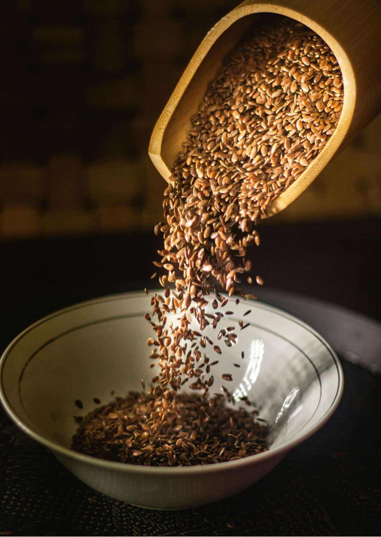 Braune Leinsamen werden aus einer Holzschaufel in eine weiße Schüssel geschüttet.