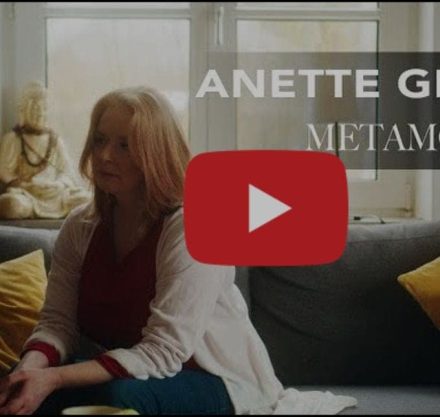 Frau mit schulterlangen blonden Haaren und weißem Hemd auf einem Sofa sitzend.