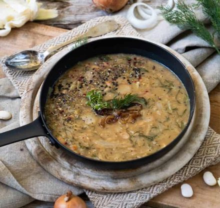 Bohnen mit gerösteten Zwiebeln in Gusseisenpfanne, garniert mit Dill und geschrotetem Pfeffer. Angerichtet auf Holzbrett mit silber Löffel und grauer Servierte.