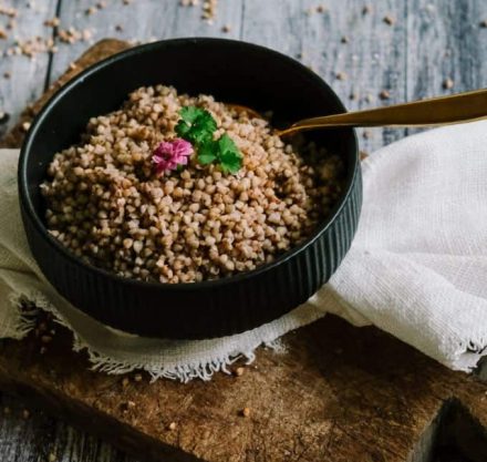 GEKOCHTER BUCKWHEAT in einer schwarzen Schüssel auf einem weißen Tuch und einem Holzbrett