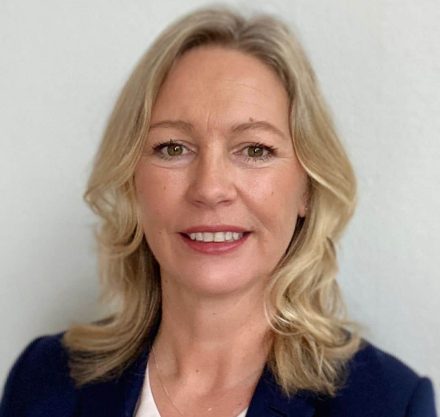 Claudia Brett mit schwarzem Mantel und weißem Hemd und blondem Haar lächelt in die Kamera
