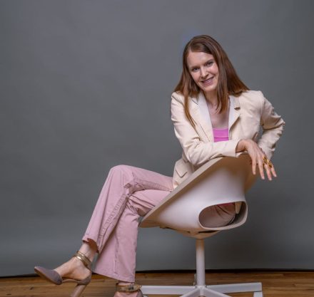 Lächelnde Frau mittleren Alters in modischer rosa Hose und beigem Blazer sitzt entspannt auf einem modernen Bürostuhl vor grauem Hintergrund
