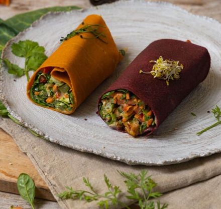 Ein roter und ein gelber Gemüsetortillas auf grauem Teller, gefüllt mit kleingeschnittenem Gemüse und garniert mit Sprossen.