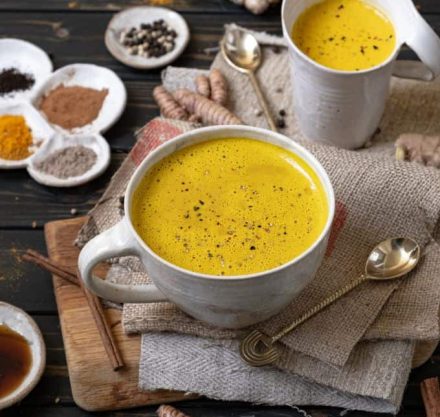 Tasse gefüllt mit goldener Milch steht auf einem mit Jutebeuteln überdecktem Holzbrett, im Hintergrund sind Gewürze auf Tellern und silberne Löffel abgebildet.