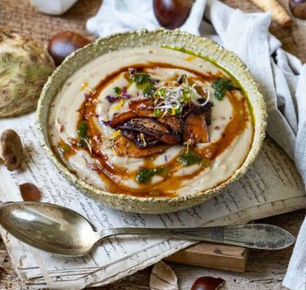 Maronensuppe in grauer Schale, garniert mit geschmorten Pilzen, Sprossen und Shoyu Sauce, angerichtet auf Holzbrett mit Silberlöffel.