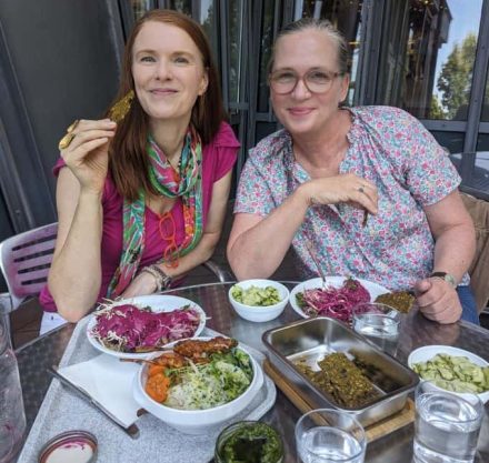 Zwei Frauen an einem mit auf weißen Tellern servierten Salat, Cracker, Dips, Gemüse gedeckten Tisch sitzend und essend.