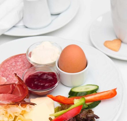 Ein klassisches Frühstück mit einem gekochten Ei, verschiedenen Käsesorten, Aufschnitt, frischem Gemüse (Paprika, Gurke, Salat), Brotaufstrichen wie Marmelade und Mayonnaise, dazu eine Tasse Kaffee mit einem kleinen herzförmigen Keks.