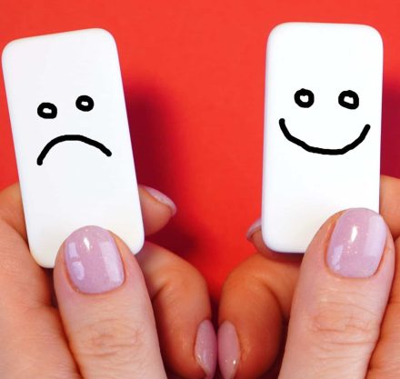 Zwei Hände mit pinkem Nagellack halten zwei weiße Kacheln nebeneinander. Auf einer Kachel ist ein handgezeichnetes trauriges Gesicht, auf der anderen ein fröhliches Gesicht.