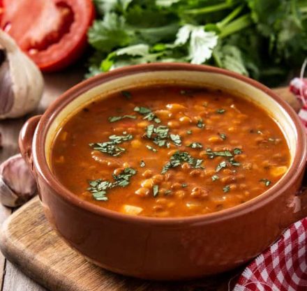 Rote Linsen- und Kirschtomatensuppe in brauner Schale auf Holzbrett serviert, mit Petersilie garniert.