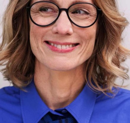Porträt einer Frau mit blonden Haaren, großer Brille und blauem Hemd