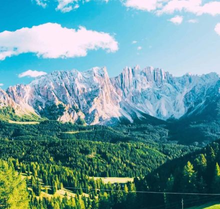 Eine malerische Landschaft mit hohen Bergen im Hintergrund, die von einem klaren blauen Himmel mit wenigen Wolken überragt werden. Die Berge haben schroffe, schneebedeckte Gipfel, während der Vordergrund von dichten Wäldern und grünen Wiesen geprägt ist. Das Sonnenlicht fällt hell auf die Szene und hebt die lebhaften Farben der Natur hervor.