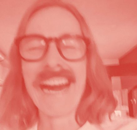 Ein verschwommenes Bild einer lachenden Person mit Brille und einem Schnurrbart. Das Bild ist in einem rötlichen Farbton gehalten, wodurch es eine künstlerische, abstrakte Wirkung hat.