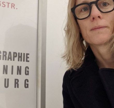 Frau mit blonden schulterlangen Haaren und schwarzer Brille und schwarzem Mantel vor einem Schild zur Mammographie.