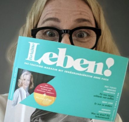Frau mit blonden Haaren und schwarzer Brille hält "Leben!" Zeitschrift vors Gesicht.