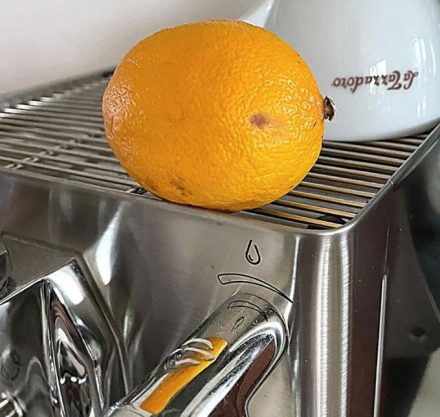 Silberne Kaffeemaschine auf der eine weiße Tasse steht und eine Zitrone liegt.