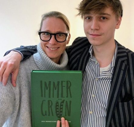 Frau im Rollenkragen Pullover und Mann im Sakko und Hemd. Beide halten ein grünes Kochbuch in der Hand.