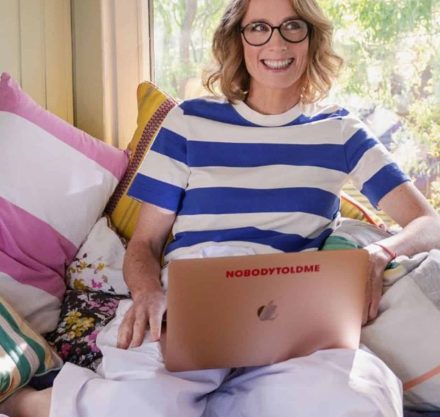 Eine lächelnde Frau mit Brille sitzt entspannt in einem sonnendurchfluteten Raum auf einem Bett voller farbenfroher Kissen. Sie trägt ein gestreiftes T-Shirt in Blau und Weiß und hat ein roségoldenes Laptop auf ihren gekreuzten Beinen, auf dessen Rückseite der Aufkleber "NOBODYTOLDME" zu sehen ist. Hinter ihr ein Fenster mit Blick auf grüne Pflanzen.