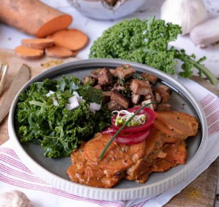 Süsskartoffel auf Grünkohl mit gebratenem Tofu angerichtet auf grauem Teller, garniert mit Tomaten und Sprossen. Platziert auf Holzbrett mit weißen Tuch.