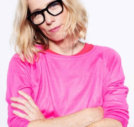 Frau in pink farbenem Oberteil und markanter schwarzer Brille hält die Arme verschrenkt vor den Körper und schaut sauer