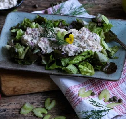 Thunfischsalat in einer blauen Schale mit violetten Blumen verziert. Der Teller steht auf einer rot-weißen Serviette und einem Holzbrett.