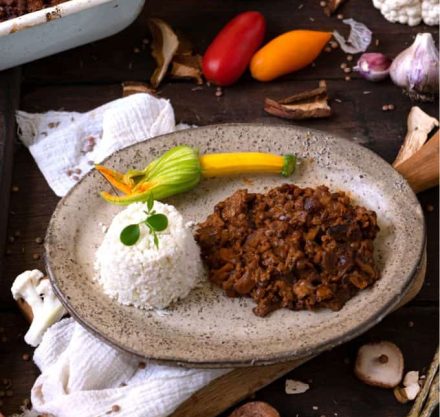 Veganes Ragu mit Blumenkohlreis auf grauem Teller angerichtet und mit Zucchini Blüte garniert.