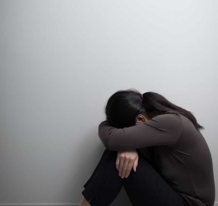 Eine Frau sitzt zusammengesunken auf dem Boden mit dem Rücken zur Wand und dem Kopf auf die Knie gelegt, was Traurigkeit und Niedergeschlagenheit ausdrückt.