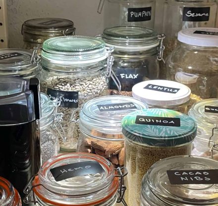 Glasgefäße in einem Vorratsschrank, ordentlich beschriftet mit verschiedenen Samen, Nüssen und Hülsenfrüchten. Zu sehen sind unter anderem Gläser mit Sonnenblumenkernen, Mandeln, Quinoa, Linsen, Kakaonibs, Hanfsamen, Leinsamen, Sesam und Kürbiskernen, beleuchtet von einer kleinen Lampe.