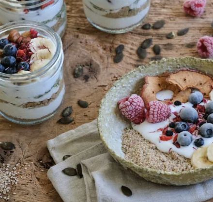 Eine Mischung aus Samen, Banane und Obst mit Joghurt auf einem Teller mit einer Serviette darunter. Es steht auf einem Holztisch