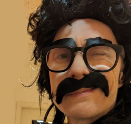 Frau mit schwarzer Kurzhaar Perücke und Brille mit angeklebtem Schnurrbart und Augenbraun