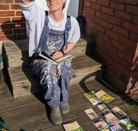 Frau in Maleranzug sitzt auf einer Holzterrasse und plant ihren Garten mit verschiedenen Gemüsesaatguttüten.