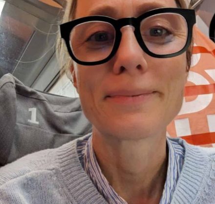 Ein Selfie einer Frau mit großen schwarzen Brillen und blonden Haaren, die einen hellblauen Pullover und eine gestreifte Bluse trägt. Sie sitzt in einem modernen Verkehrsmittel, vermutlich einem Zug, auf einem Sitzplatz mit der Nummer "1".