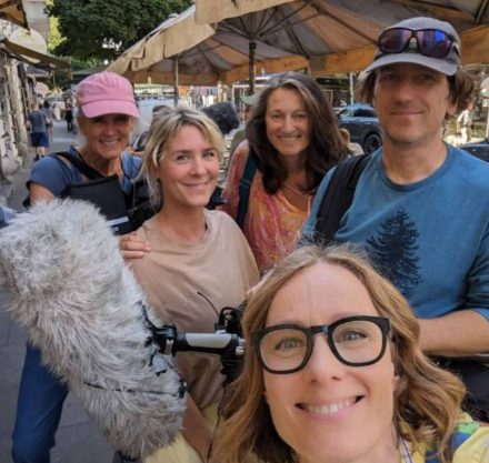 Eine Gruppe von fünf Menschen, die fröhlich in die Kamera lächeln. Die Person im Vordergrund trägt eine Brille und ein gelb-weiß gestreiftes Hemd. Eine andere Person hält ein Mikrofon mit einer Fellabdeckung. Sie stehen auf einer belebten Straße unter Sonnenschirmen, vermutlich in einem städtischen Bereich mit Cafés und Geschäften im Hintergrund.