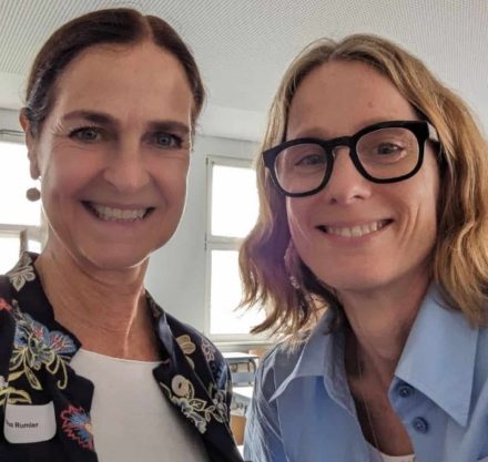 Ein Selfie von zwei Frauen, die lächelnd in die Kamera schauen. Die Frau links trägt eine Jacke mit buntem Blumenmuster und ein Namensschild mit der Aufschrift "Andrea Rumler". Die Frau rechts trägt eine Brille und ein hellblaues Hemd. Im Hintergrund befindet sich ein heller Raum mit Fenstern und Tischen.