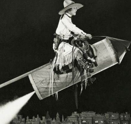 Schwarz- weiß Foto eines Cowboys auf einer Rakete über eine Stadt fliegend