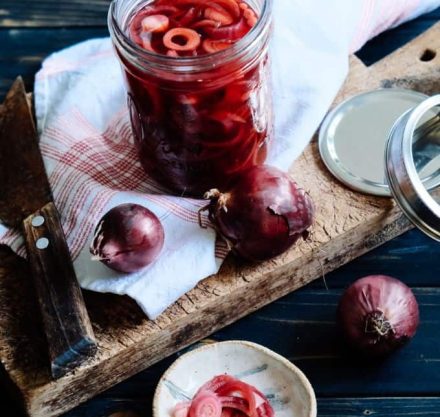 Eingelegte rote Zwiebeln in einem Glasbehälter auf einem hölzernen Schneidebrett, während rohe Zwiebeln den Behälter umgeben