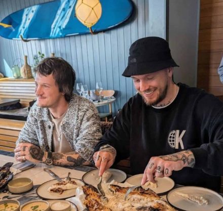 Zwei lächelnde Männer sitzen an einem Tisch in einem Restaurant mit maritimem Flair, erkennbar an der blauen Wand und einem Surfbrett als Dekoration. Beide haben sichtbare Tätowierungen und der eine trägt eine schwarze Mütze. Vor ihnen auf dem Tisch liegt ein gegrillter Fisch, an dem einer der Männer gerade schneidet, umgeben von Tellern und Saucenschälchen, bereit zum gemeinsamen Essen.